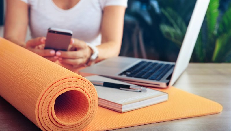A half-rolled yoga mat lies on a table with a notebook, pen and laptop, while a woman types on a mobile phone in the background
