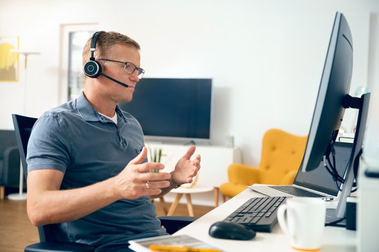 A man wearing glasses, a polo shirt, and a headset gesticulating as he sits in front of a home workstation