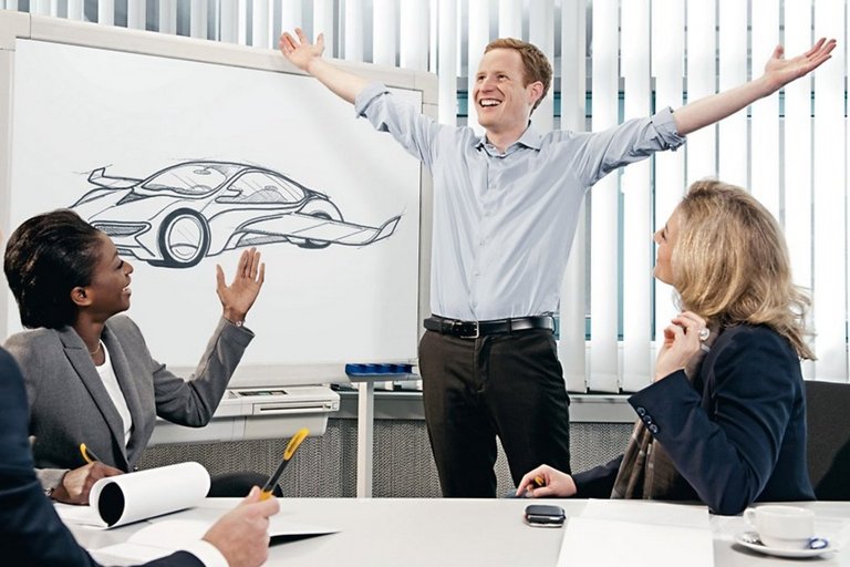 A man stands in front of a whiteboard depicting a car with wings and stretches both arms happily from himself while two women and a man look at him