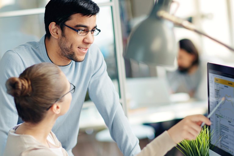 A man with glasses is standing at a desk looking at what a woman with glasses is showing him sitting on a display