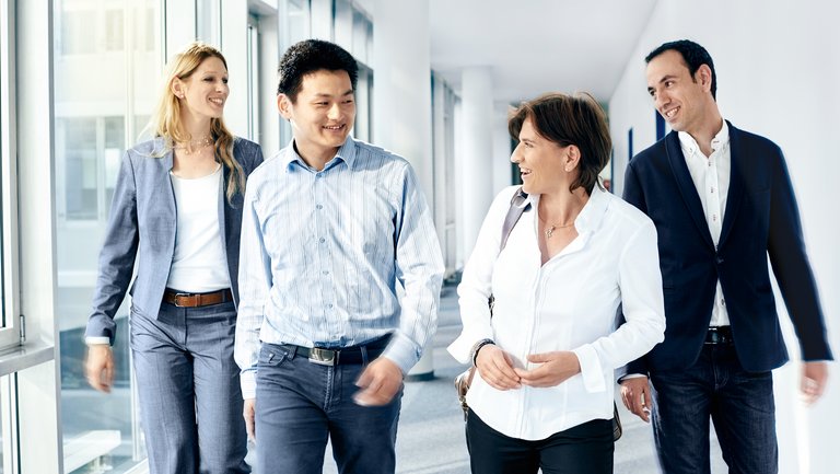 Two men and two women in business outfits walk down a light-flooded corridor with floor-to-ceiling window panes and smile at each other