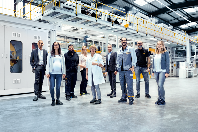A group of people from different ethnic backgrounds and with different job positions stand in a factory hall