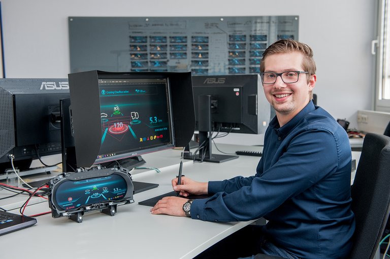 A man is sitting in front of a computer monitor next to an instrument cluster and is working with a graphic tablet