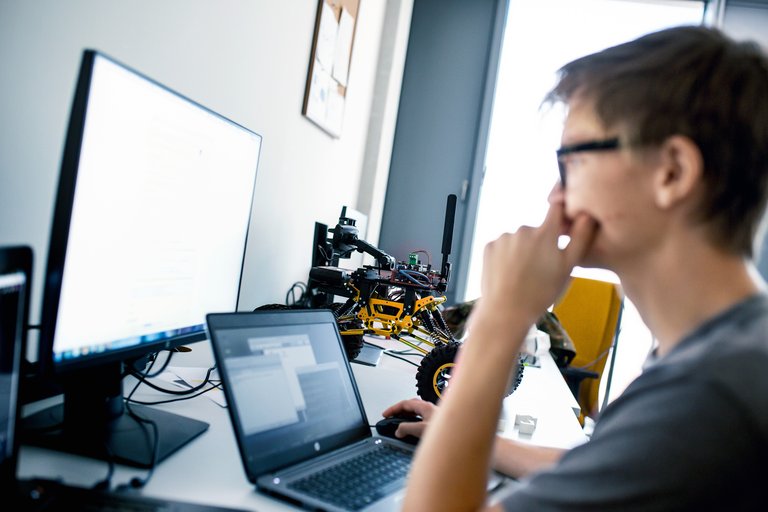 A young man with glasses sits in front of a laptop and a screen while a toy racing car stands in the background